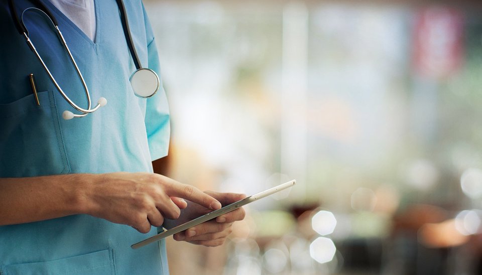 Half body image of medical staff in blue scrubs inputting data into a tablet