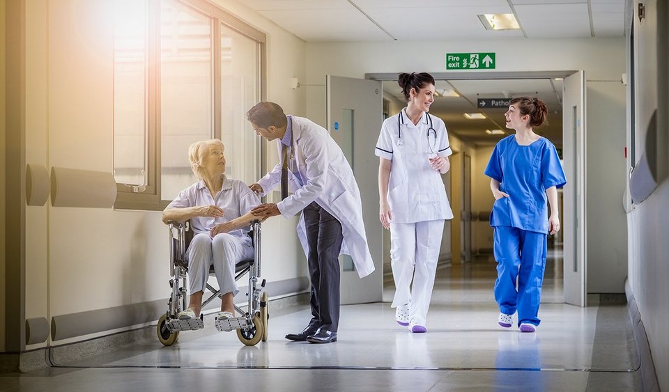 Hospital corridor where a male consultant is speaking to a patient in a wheel chair and two female medical staff are following behind them